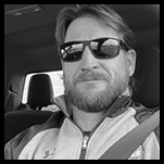 Mike McGowan of Sales, with sunglasses and a beard, sitting in a car's back seat.