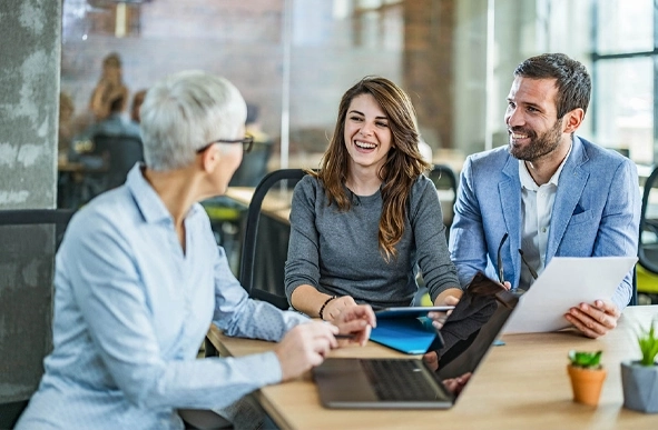 Three professionals discussing work at a table, focused on a laptop screen.